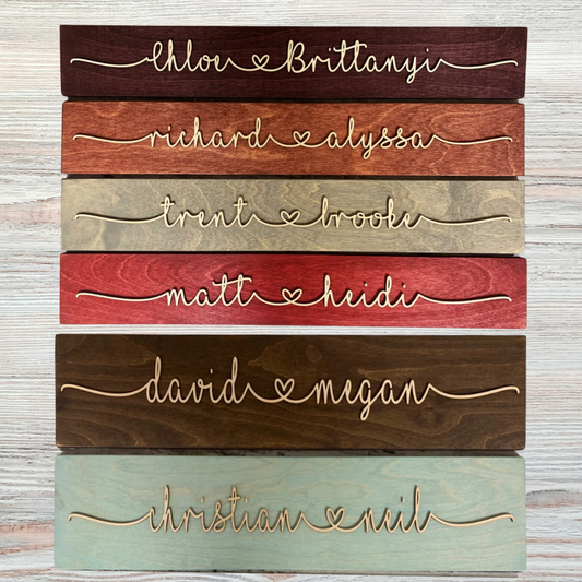Six horizontal wood plank signs featuring pairs of laser-cut names connected by a heart. Each sign has a different stained background, including burgundy, red, brown, gray, teal, and natural wood tones. The names are written in a flowing script with a heart in the center, mounted on a light-colored wood background