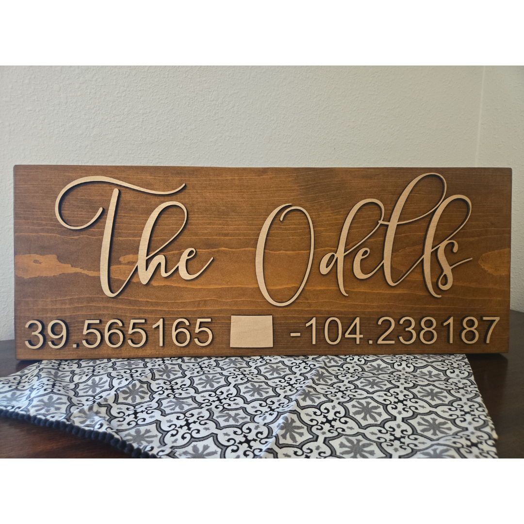 A wooden plank sign with the text 'THE ODELLS' laser cut on top, followed by a set of coordinates.