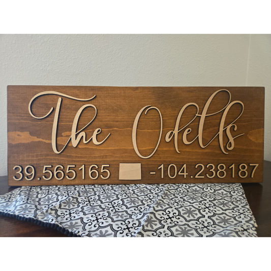 A wooden plank sign with the text 'THE ODELLS' laser cut on top, followed by a set of coordinates.