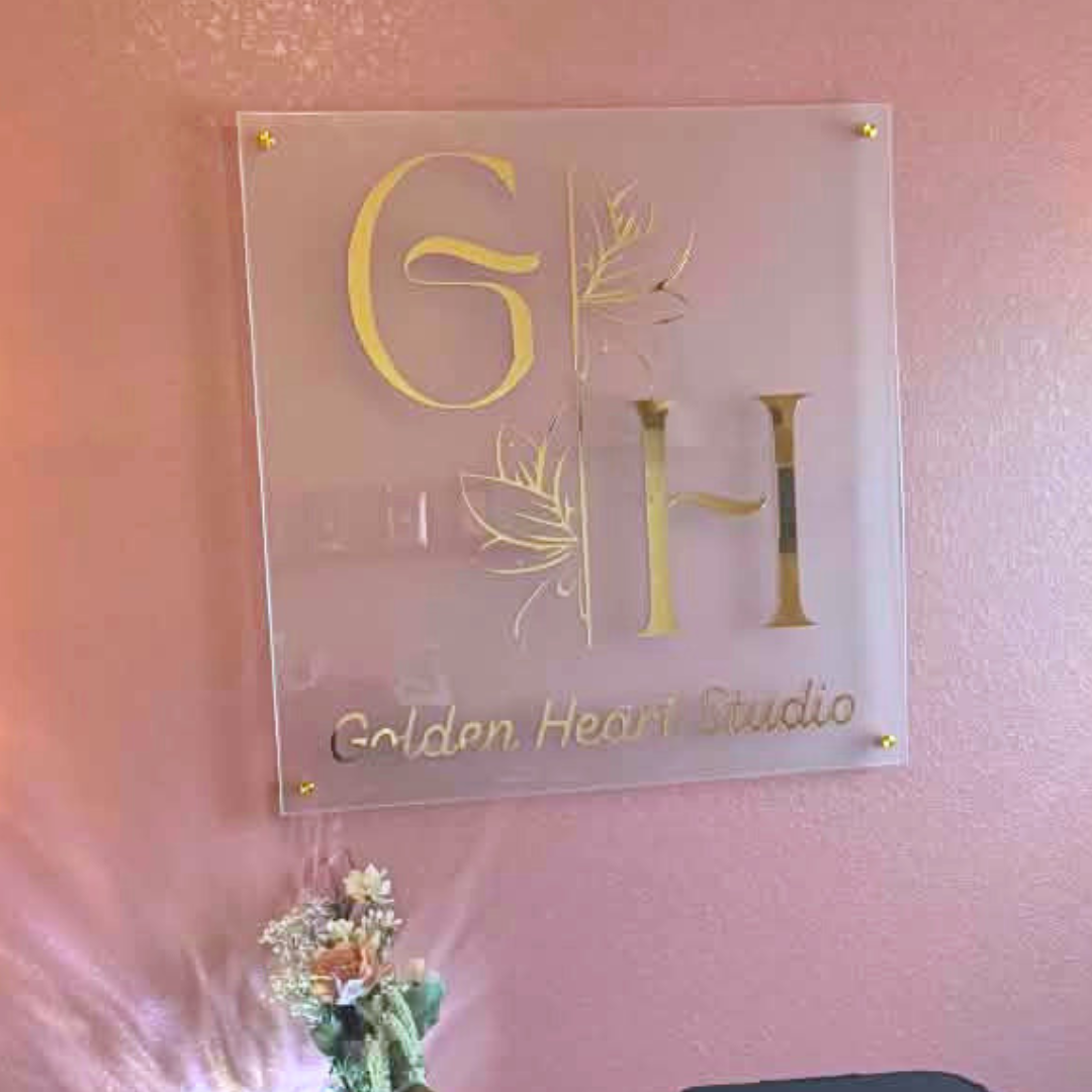 A square acrylic wall sign with gold lettering reading 'GH' and 'Golden Heart Studio,' mounted on a pink wall above a small vase of flowers and a dark bag.
