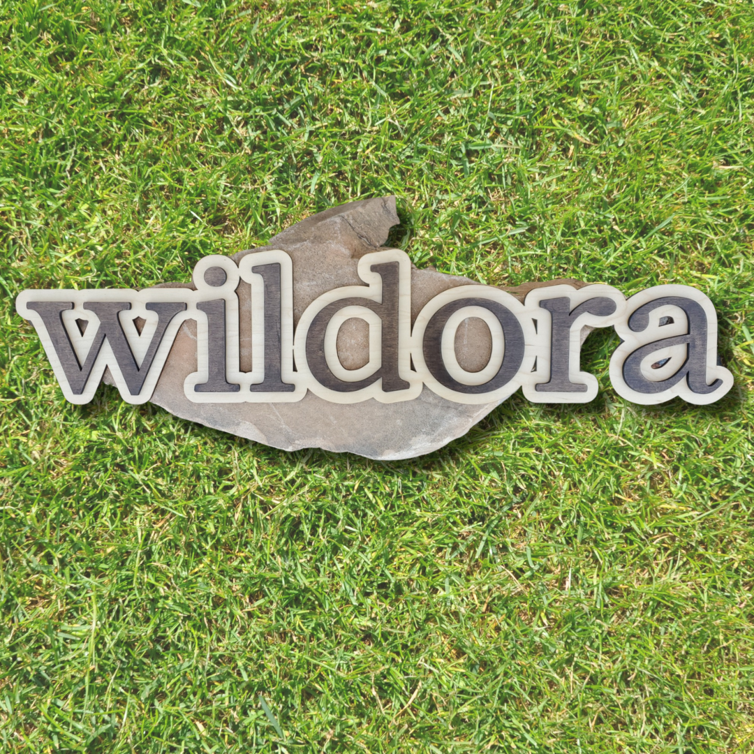 A custom wooden sign with the word 'wildora' layered in different shades of brown, placed on a grass background.
