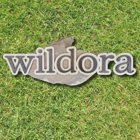 A custom wooden sign with the word 'wildora' layered in different shades of brown, placed on a grass background.