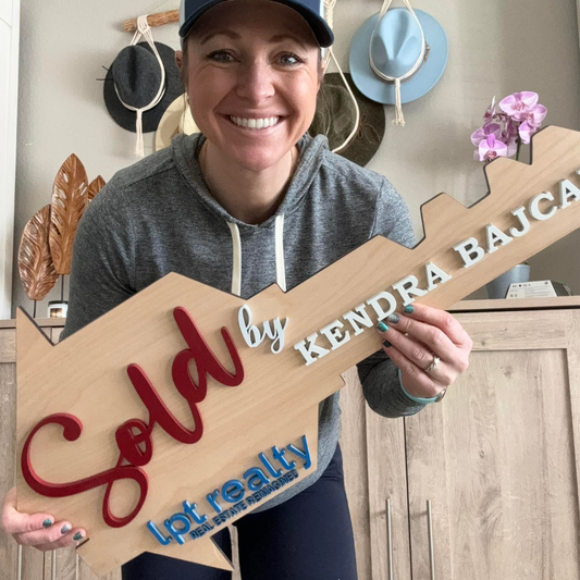 A smiling woman holding a wooden real estate sign with the text 'SOLD by Kendra Bajcar', with a background of various hats and a potted plant.