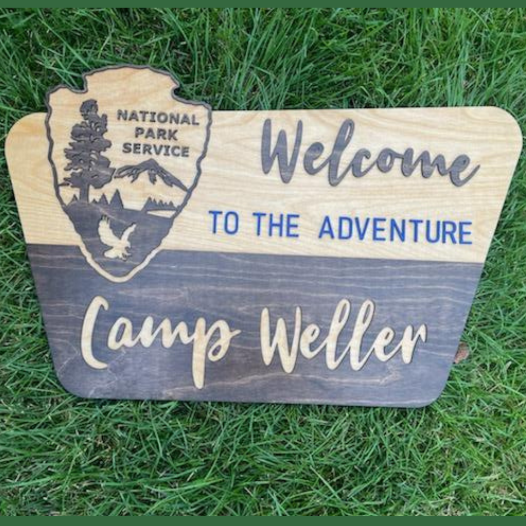 A wooden sign with the National Park Service emblem and the text 'Welcome to the Adventure' in bold lettering, placed on grass.