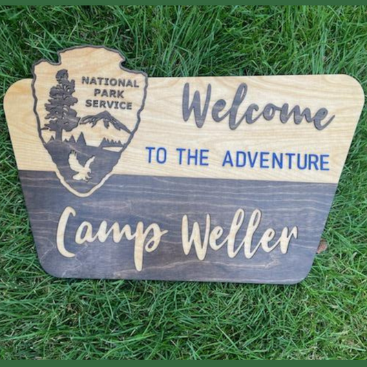 A wooden sign with the National Park Service emblem and the text 'Welcome to the Adventure' in bold lettering, placed on grass.
